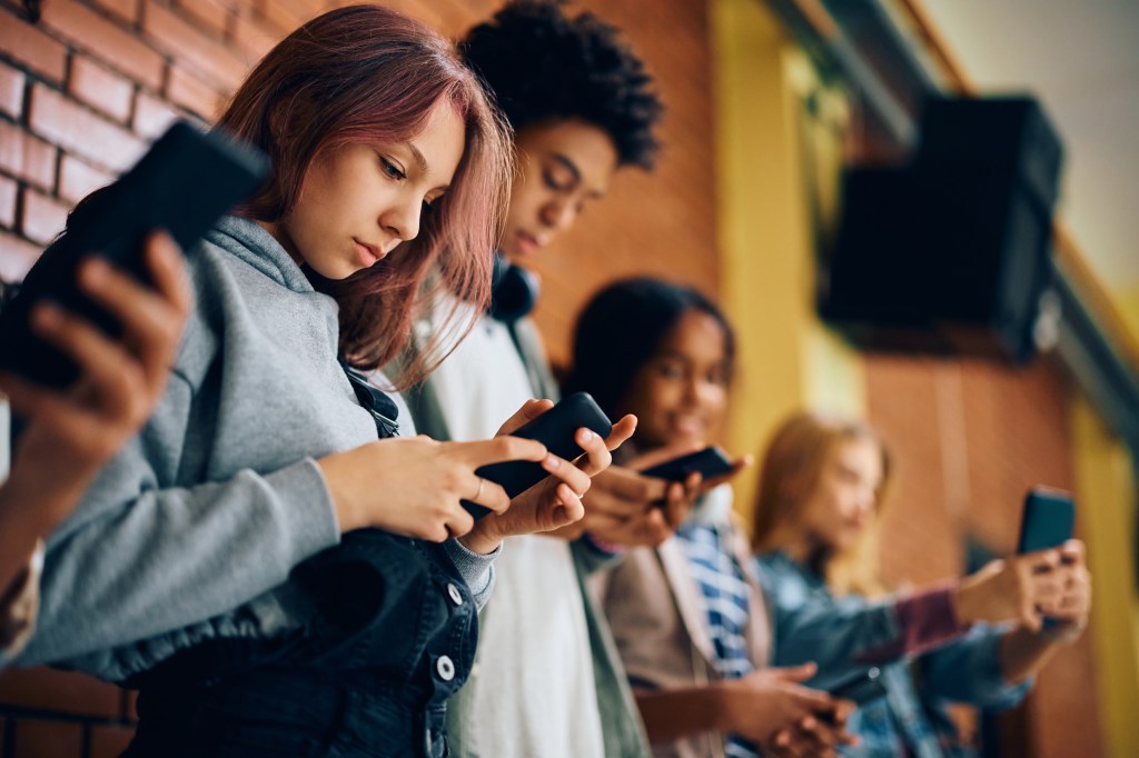 A group of diverse teenagers uses smartphones in a school hallway.