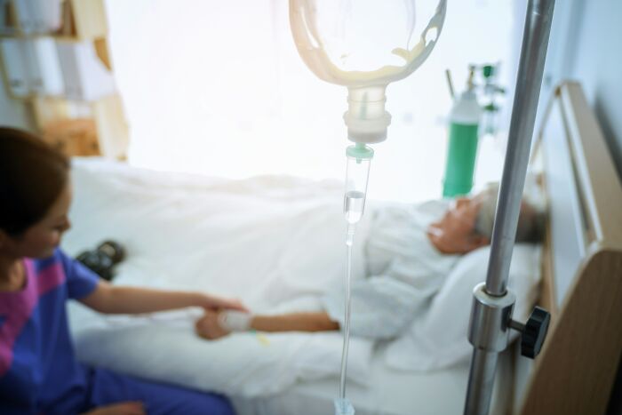 An IV drip in focus with a nurse attending to a patient in the background, depicting hospital horror stories.