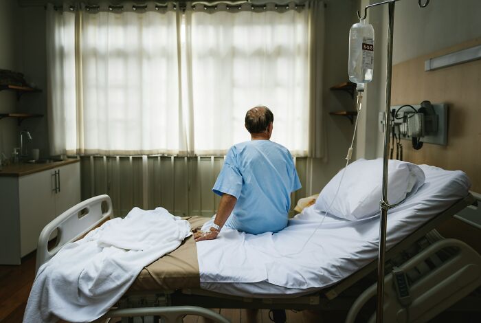 A patient in a hospital gown sits on a bed, facing away, highlighting the isolation in hospital horror stories.