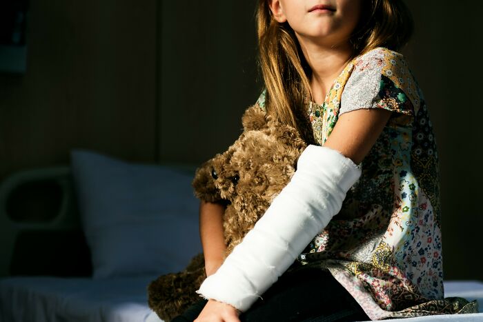A young girl with a cast on her arm, holding a teddy bear, illustrating hospital horror stories.