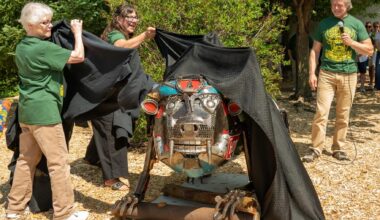 Three adults unveil a metal animal sculpture outdoors, pulling back a black cloth. Trees and wood chips are visible around them.