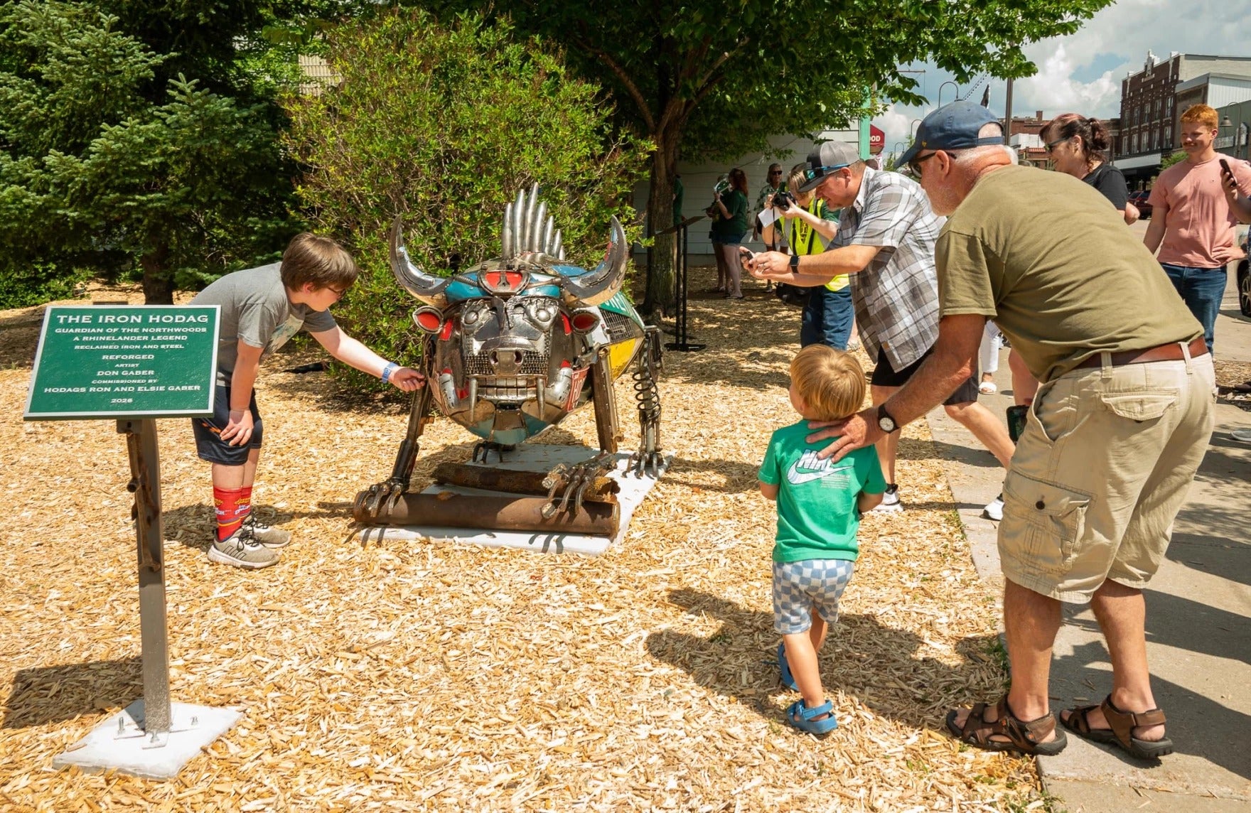 Adults and children view and photograph a metal sculpture called The Iron Hodag outdoors, with a sign describing the artwork on the left.
