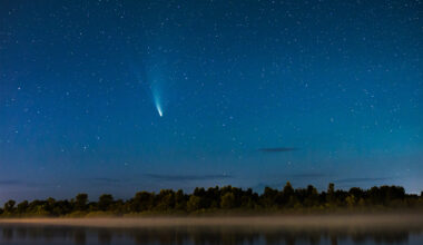 A bright-tailed comet streaks across a starry night sky above a fog-covered river and treeline.