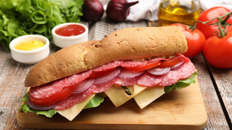 A sub sandwich layered with cold cuts, tomato, onion, cheese, and lettuce sits on a wooden cutting board