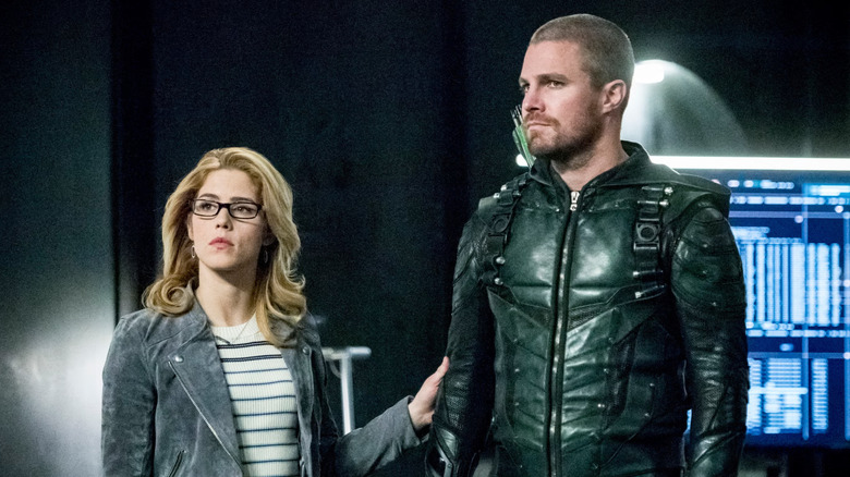 Oliver Queen suited up as the Green Arrow, sans mask, next to Felicity Smoak in the Arrowcave on Arrow.