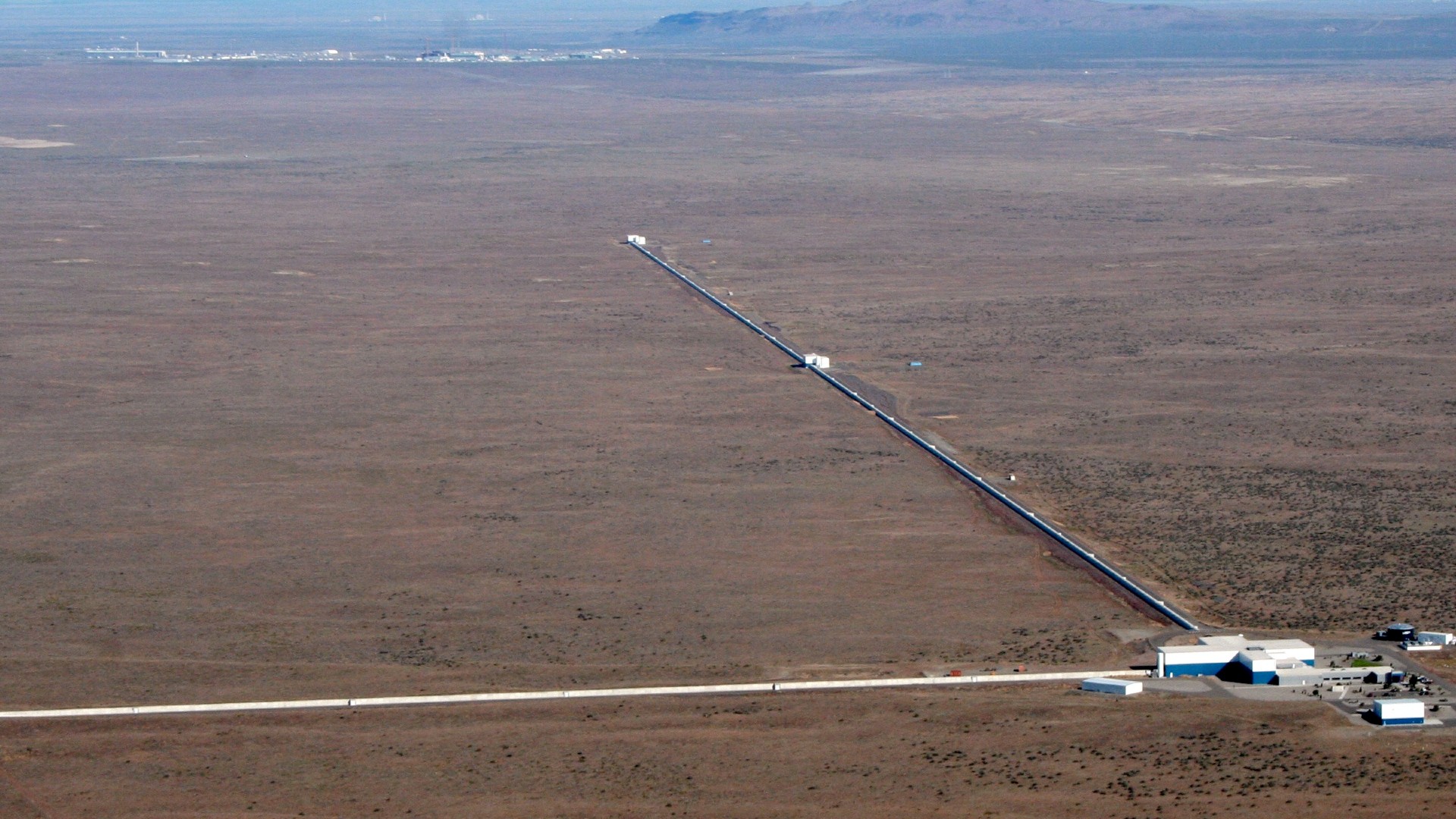 An aerial shot of a brown plain. There is a white building toward the bottom left and two extremely long arms shoot out from the building, forming a 90 degree angle.