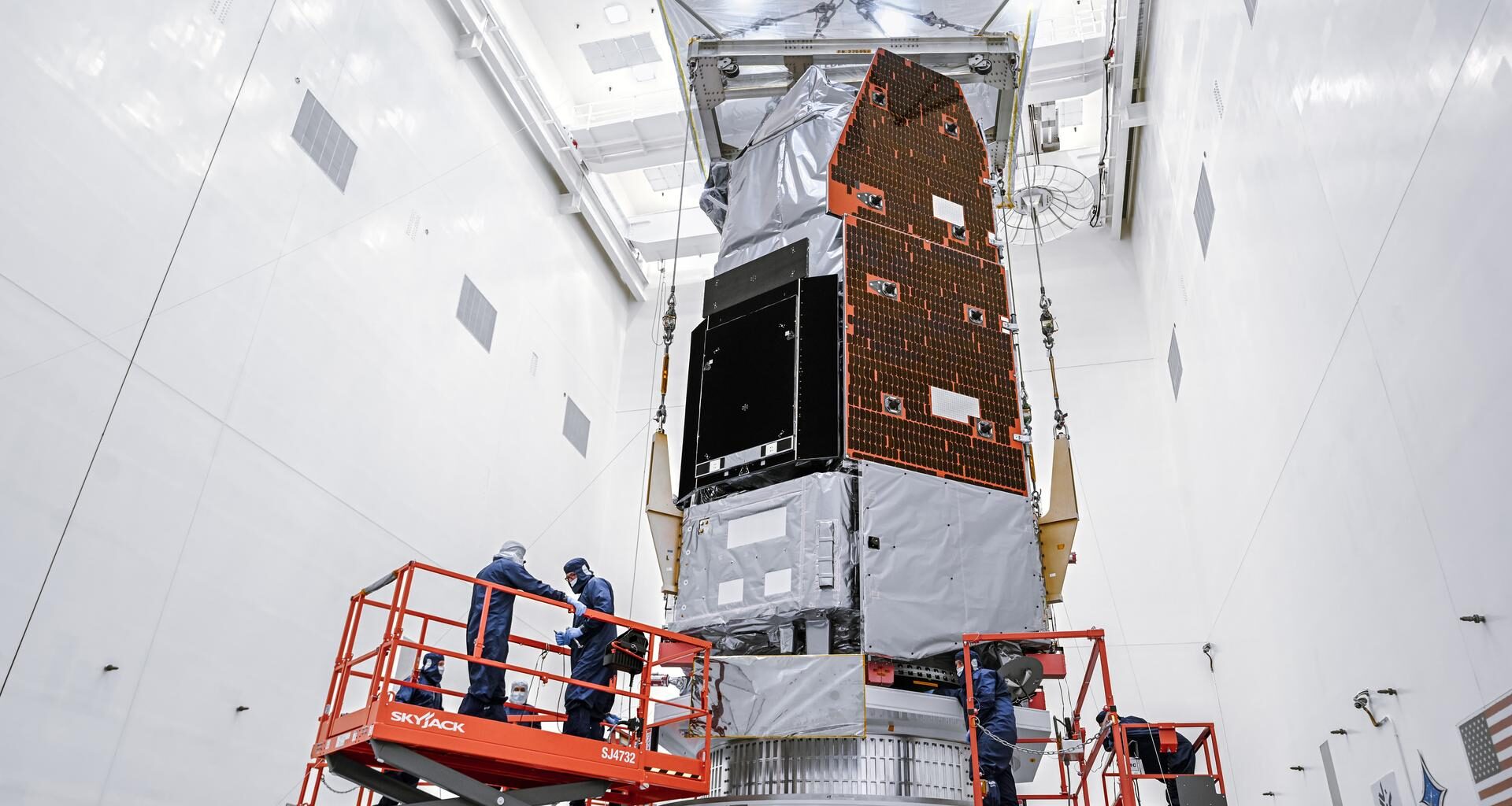 A large silver and gray space telescope sits in a brightly lit, white clean room. Team members in blue suits work on the telescope from orange mechanical lifts.