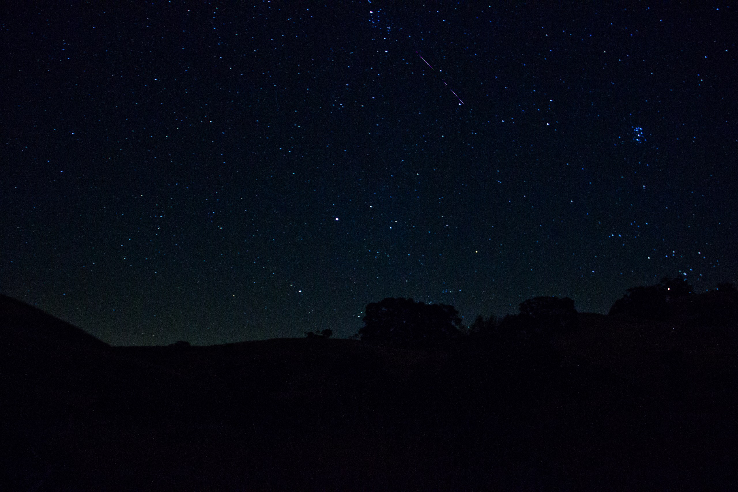 A wide-angle night sky photograph showing a lone meteor streaking diagonally across the upper portion of the frame against a deep blue, star-filled sky. The Pleiades star cluster is visible as a tight grouping of blue-white stars in the upper right. The foreground is a dark silhouette of rolling hills and trees, with a faint green glow of airglow near the horizon.