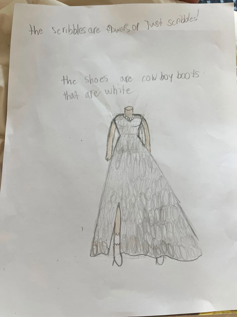 A child's pencil drawing of a long wedding dress with a fitted bodice, long sleeves and a full skirt. Notes explain that the scribbles are "flowers or just scribbles" and that the bride is wearing white cowboy boots.