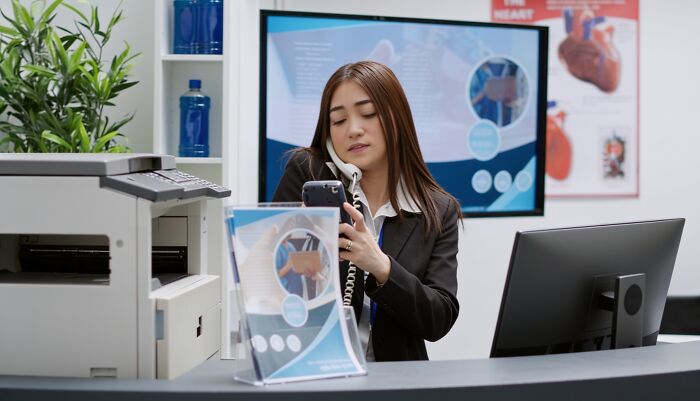 A busy medical industry receptionist answers two phones simultaneously at a front desk with medical information displayed.