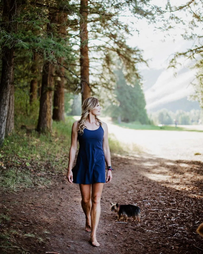 Mariel Hemingway walking barefoot on a forest path in a navy dress with her small dog, sunlight streaming through the trees.