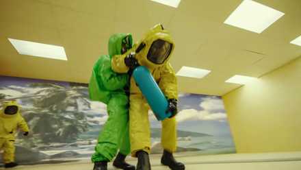 A figure in a green Hazmat suit clings to someone in a green one as they stand in a yellow-wallpapered room in Backrooms