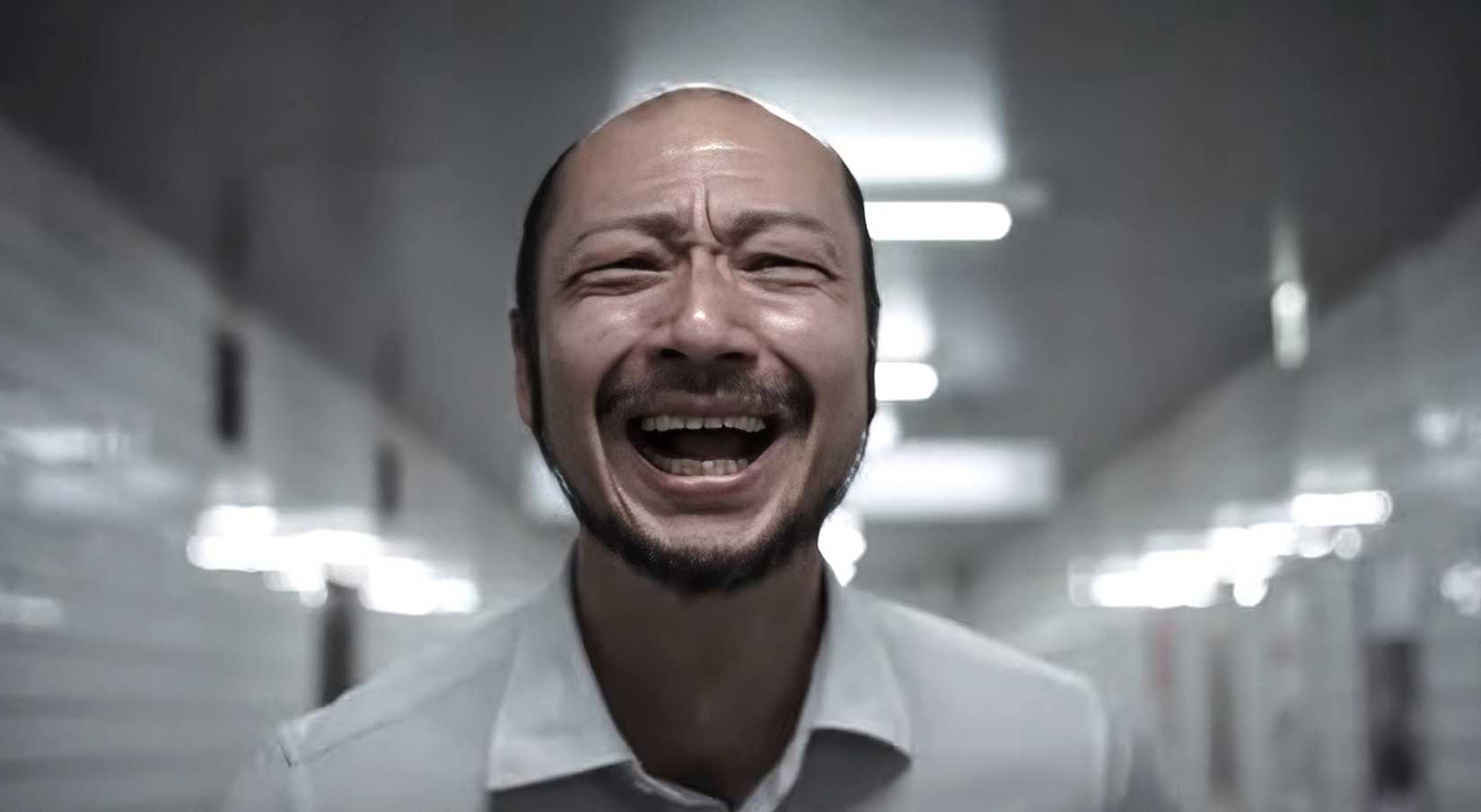 A close-up of a thin, balding Japanese man laughing with a rictus grimace that seems like pain in Exit 8, the movie