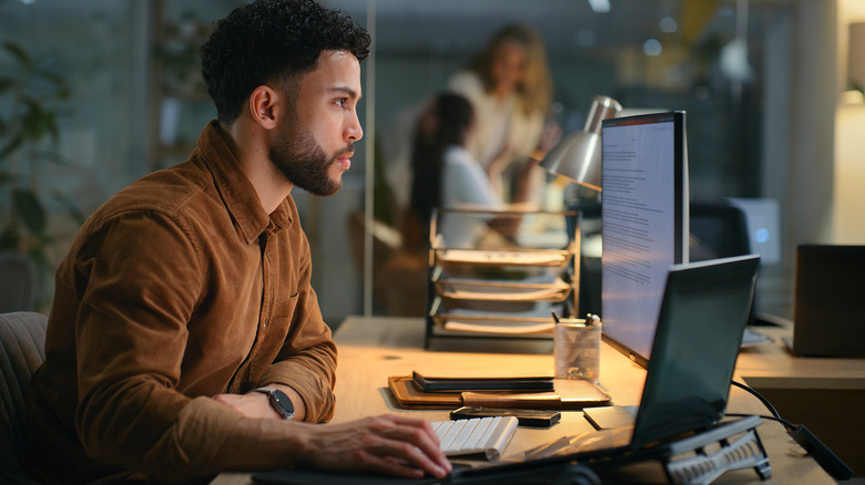 A man in a brown shirt working at a multi-screen desk setup in a warmly lit office.