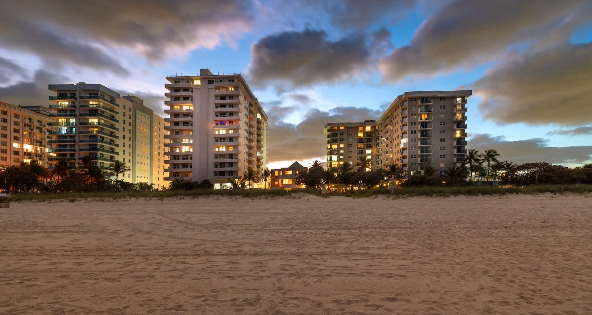 A beach view at dusk featuring tall buildings with illuminated windows and dramatic clouds in the sky.