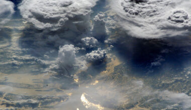 Towering storm clouds over part of Earth’s surface are seen from space, with sunlight reflecting off part of the ocean’s surface.