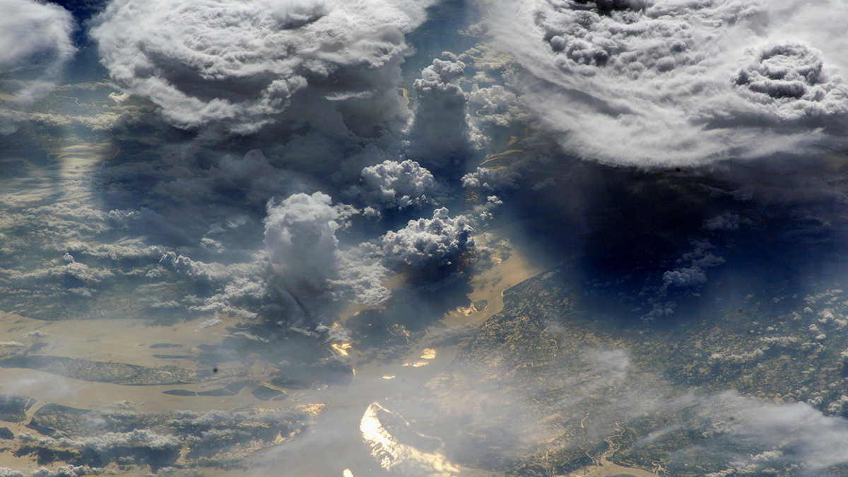 Towering storm clouds over part of Earth’s surface are seen from space, with sunlight reflecting off part of the ocean’s surface.