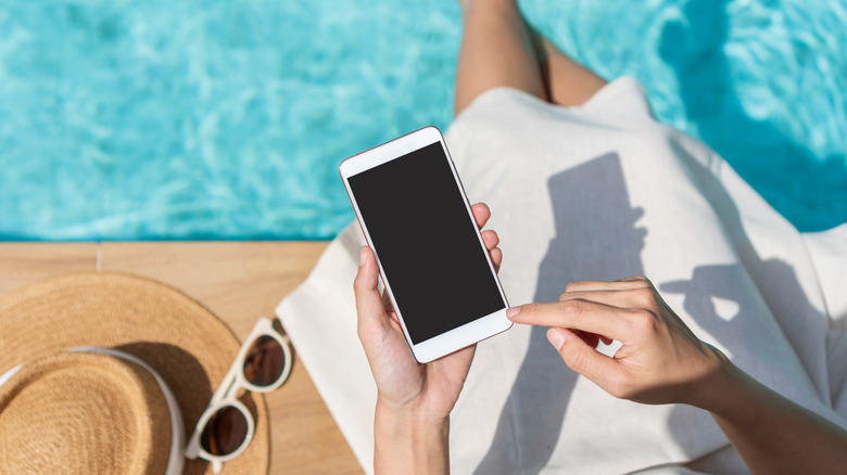 A person using a phone while sitting beside a swimming pool, on a sunny day.