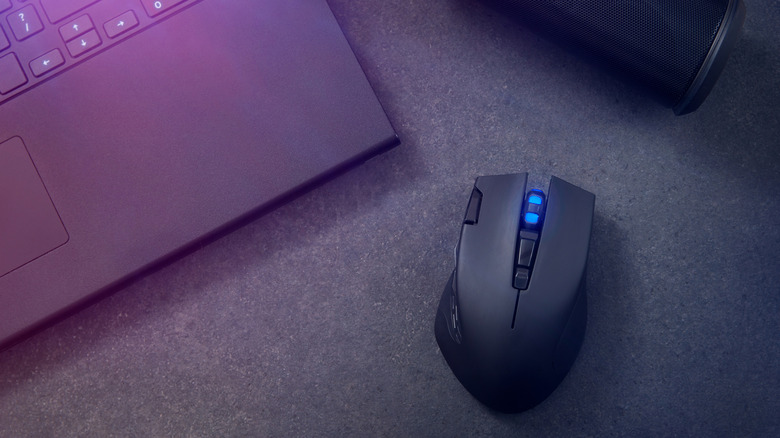 A top-down view of a black wireless multi-button mouse with a glowing blue scroll wheel sitting next to a laptop and a cylindrical speaker.