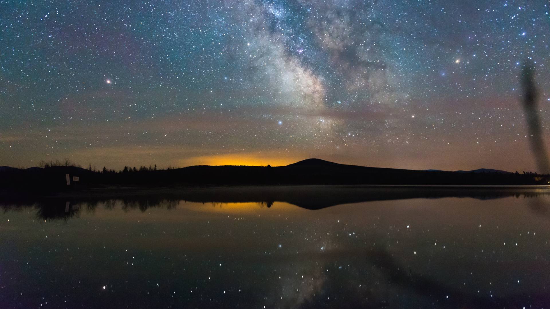A star-filled night sky with the Milky Way visible, reflected over a still lake, with distant hills silhouetted against a faint orange glow on the horizon.