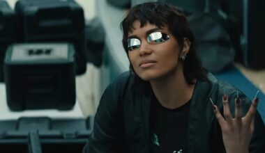 girl with short cropped dark hair wearing futuristic glasses and holding up a hand with long metallic fingernails