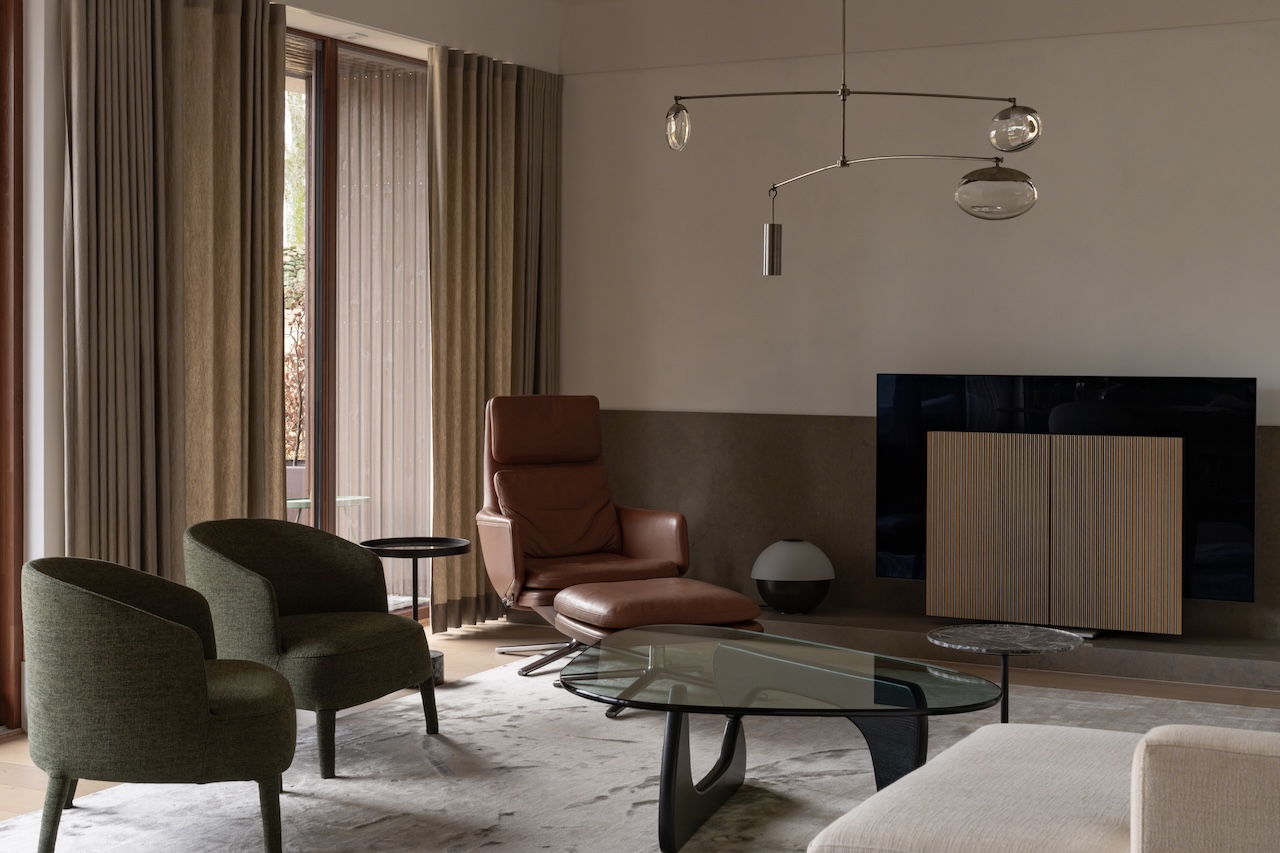 Modern living room with green armchairs, brown leather recliner, glass coffee table, TV, and large windows with beige curtains. Neutral color palette and minimalist decor.