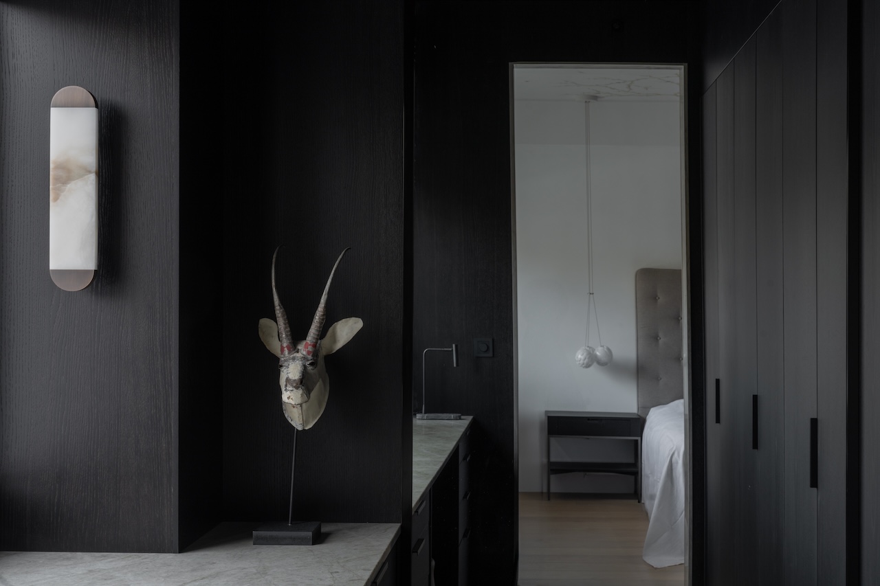 A dark modern hallway with a mounted antelope skull leads into a minimalist bedroom with a tufted headboard and hanging bedside light.