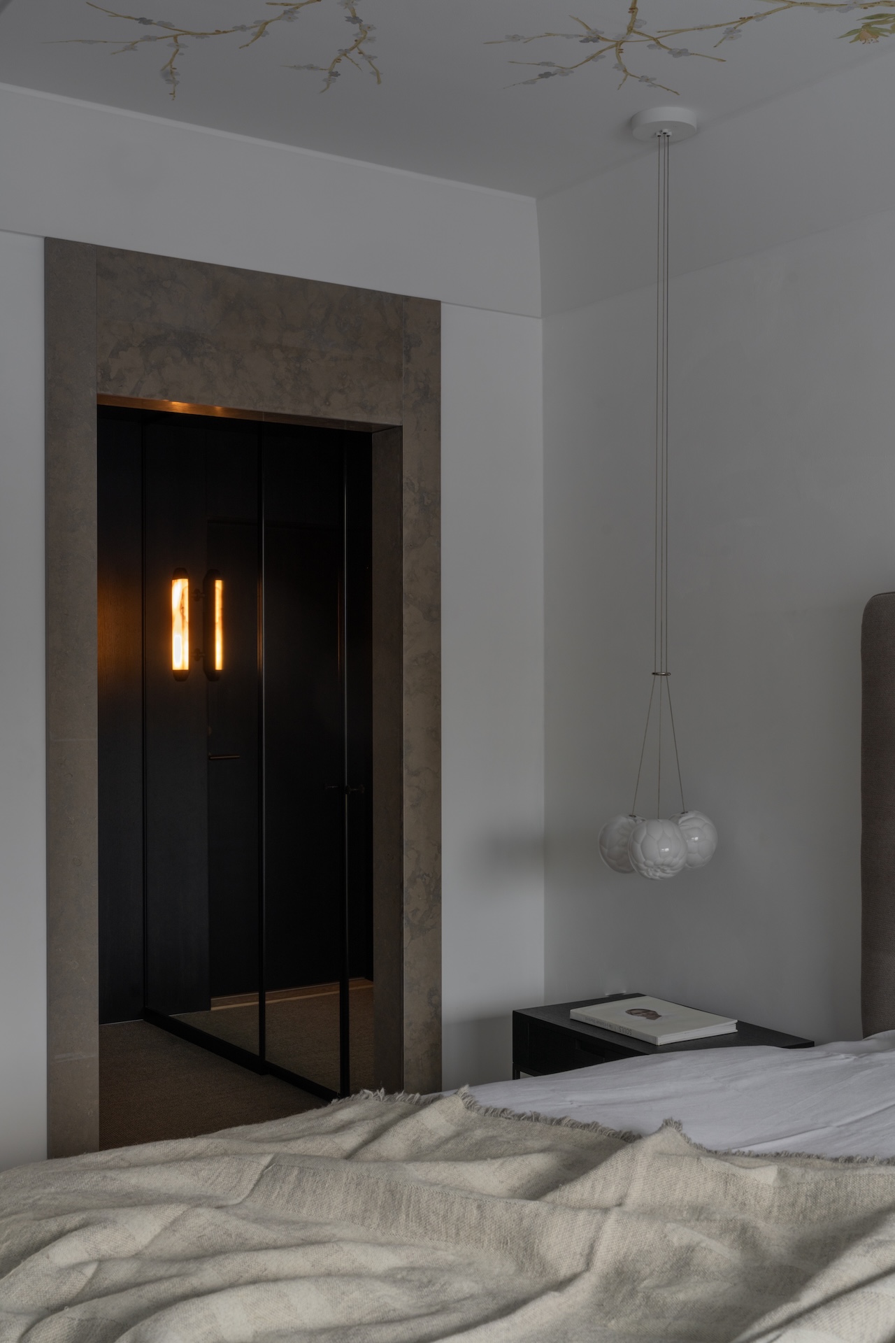 Minimalist bedroom with a beige blanket on the bed, a dark nightstand with a book, modern pendant light, and a doorway showing a lit wall sconce in a dark hallway.