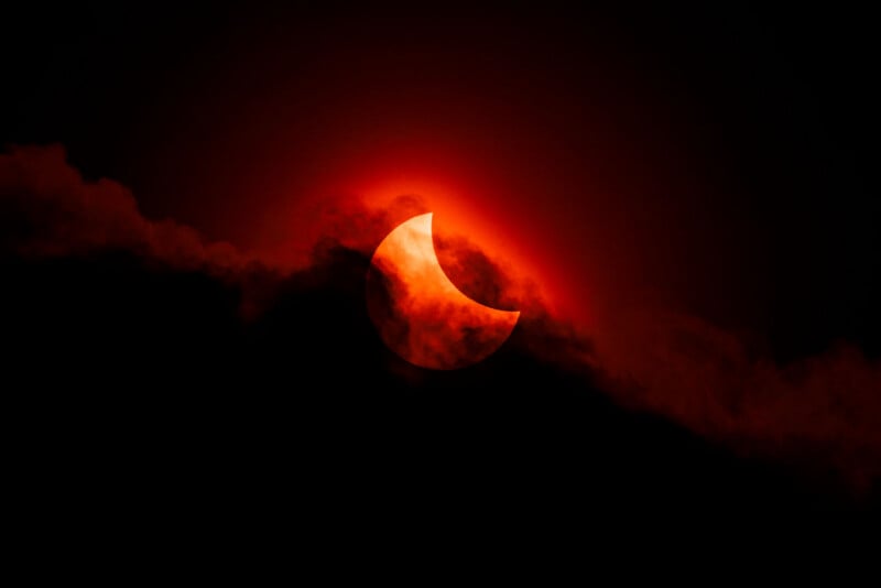 A partial solar eclipse with the sun partially obscured by the moon, glowing red and orange behind dark clouds in a dramatic night sky.