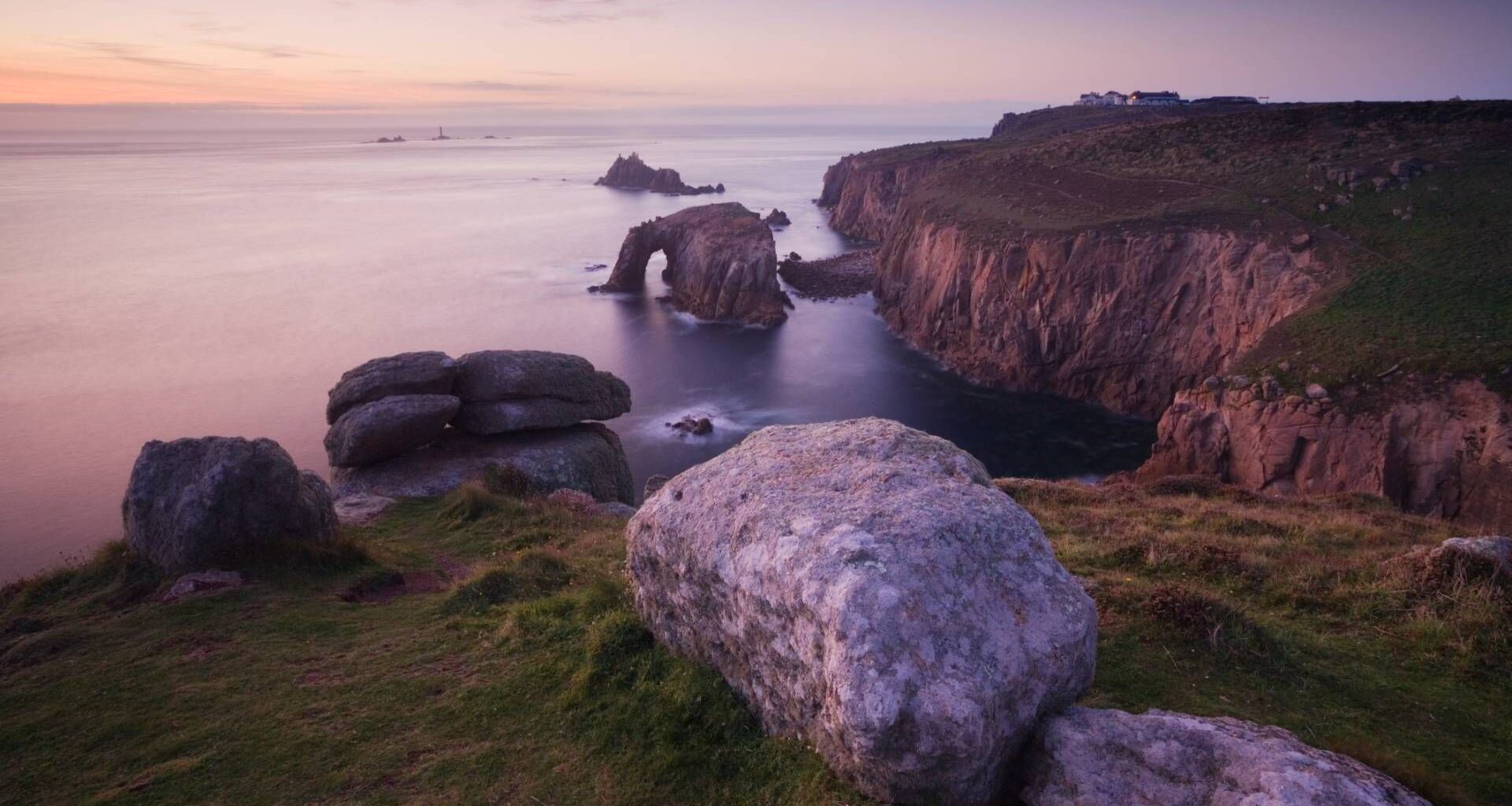 Rocky coastal cliffs (Land's End, Cornwall, UK) at sunset with a natural stone arch extending into the sea, grassy foreground, and calm water. In the far distance is a lighthouse.