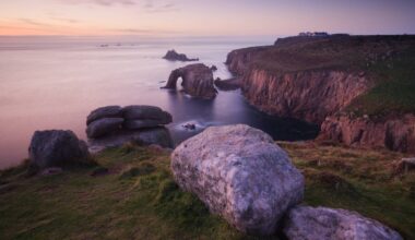 Rocky coastal cliffs (Land's End, Cornwall, UK) at sunset with a natural stone arch extending into the sea, grassy foreground, and calm water. In the far distance is a lighthouse.
