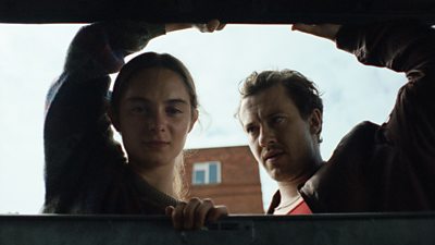 A scene from Hoard. Two people look inside a rubbish bin. One character watches the other with a concerned expression. 