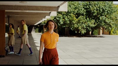 A scene from Club Zero. A woman in a bright orange t-shirt walks across a concrete plaza. Teenagers in school uniforms are in the background.