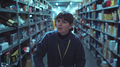 A scene from On Falling. A woman walks down an aisle in a large storage facility. Documents and items are piled on shelves on either side of her. 