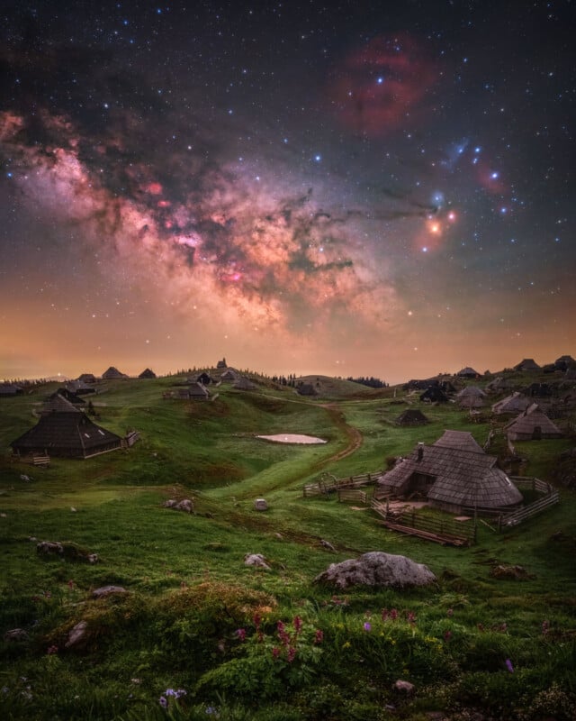 A vibrant night sky full of stars and the Milky Way stretches over green rolling hills dotted with rustic wooden huts, fences, and a small pond, with wildflowers in the foreground.