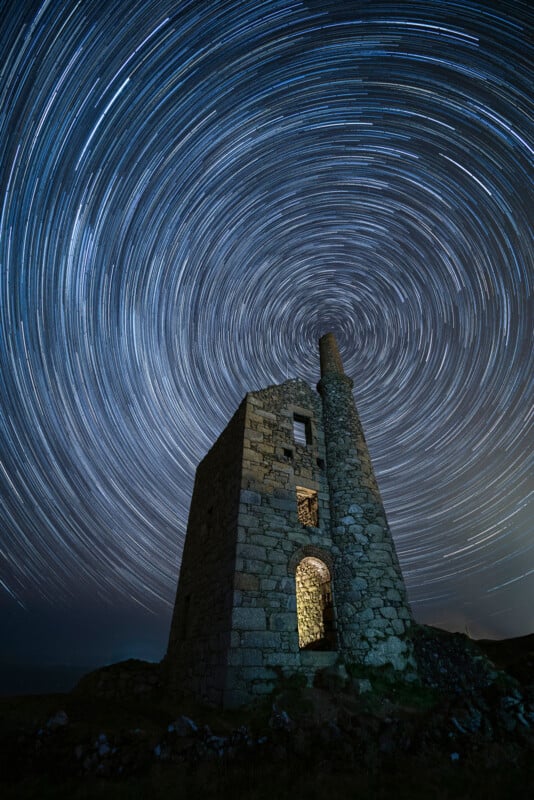 A long-exposure photo of a stone tower at night, with swirling star trails creating circular patterns in the sky above the structure. Light illuminates the tower’s arched doorway and some windows.