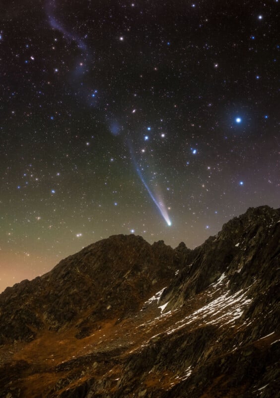 A bright comet with a glowing tail streaks across a star-filled night sky above rugged, rocky mountains with patches of snow.