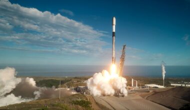 A white, black and gray-ish (soot-covered) rocket lifts off into a mostly-blue sky from a sea-side launch pad.