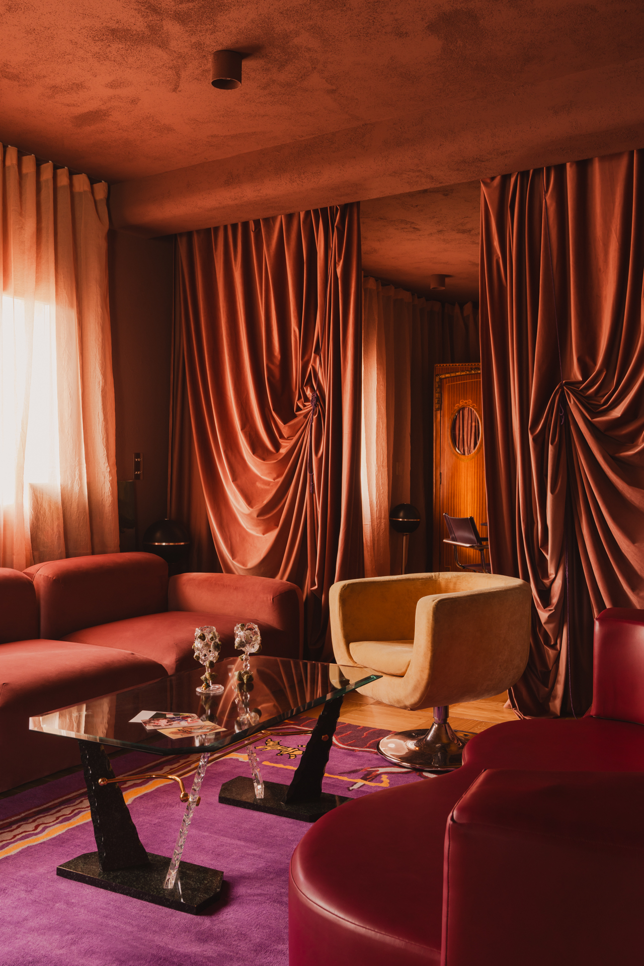 A living room with a glass coffee table and mixed seating upholstered in red leather and velvet, all backed by a doorway draped in fabric.