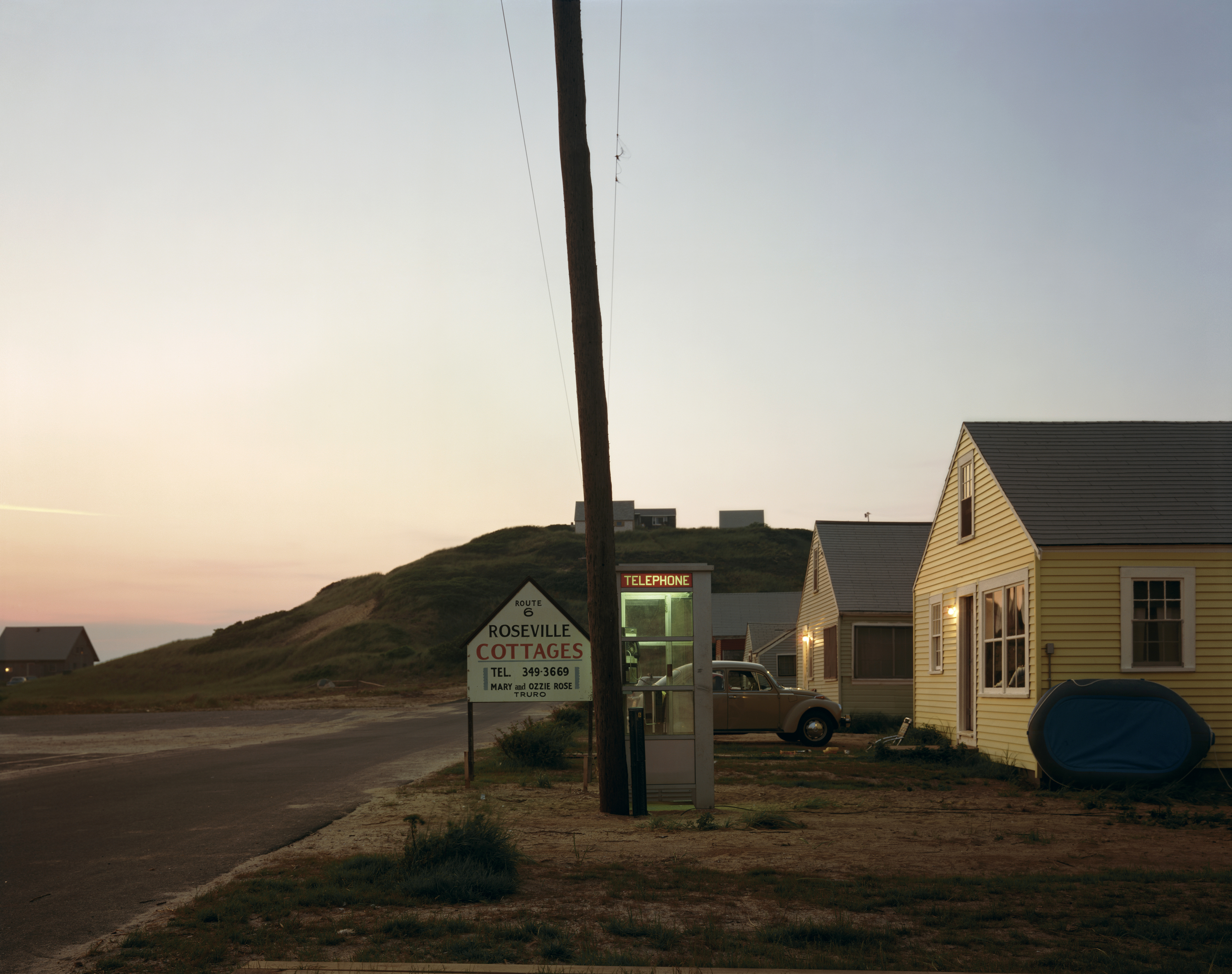 At dusk, a lit telephone box stands beside a hand-painted Roseville Cottages sign and a row of yellow shingled cabins, with a vintage car parked nearby.