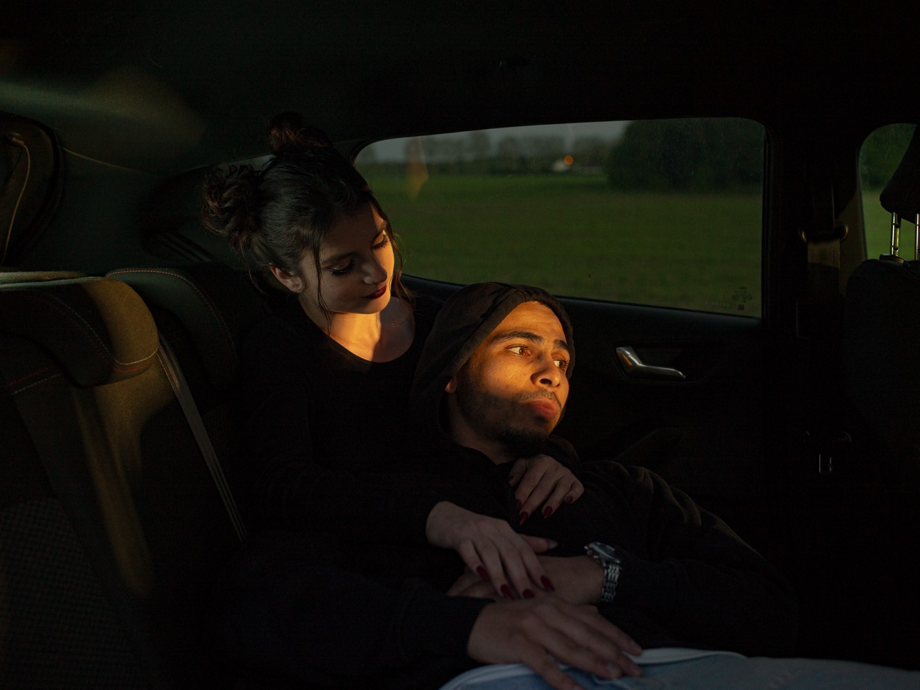 A young man and woman sit closely in the backseat of a car, illuminated by warm, low sunlight shining through the window as dusk settles over the fields outside.