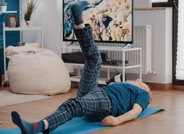 Elder man laying on yoga mat and training muscles, lifting legs to do physical exercise and workout. Senior person doing fitness activity with sport equipment at home. Adult stretching