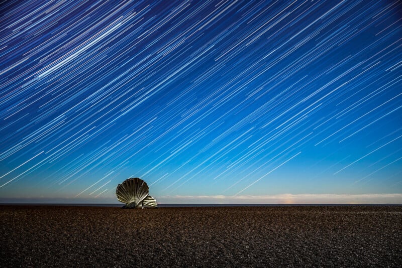 A large shell-shaped metal sculpture stands on a pebble beach under a night sky filled with bright star trails, creating streaks of light across the deep blue sky.