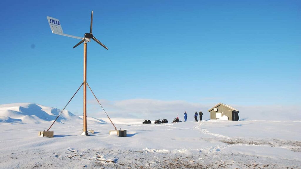 Wind turbine on snowy landscape with a small building and people in the background.