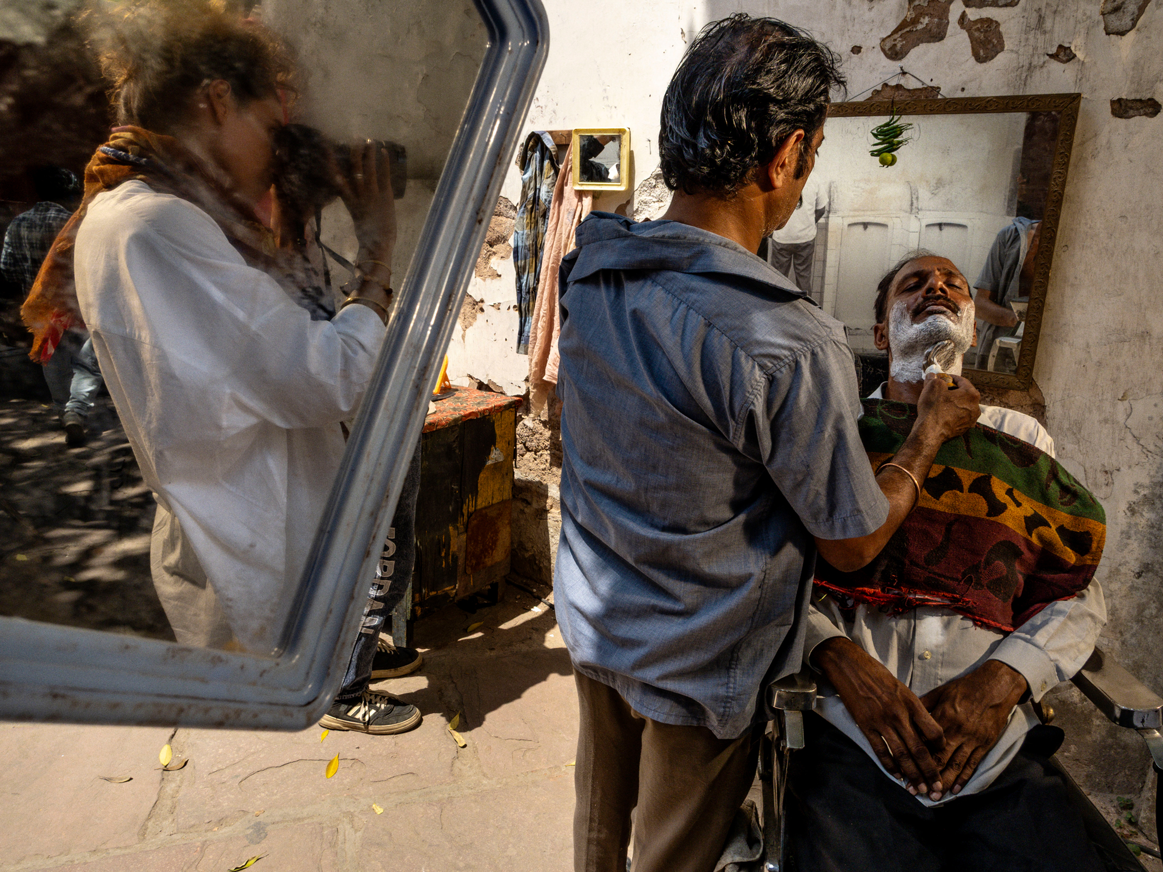 An image that on the right shows two men in a backyard in India, with one standing in front of the other and shaving him. On the right is a mirror that shows the reflection of a female photographer