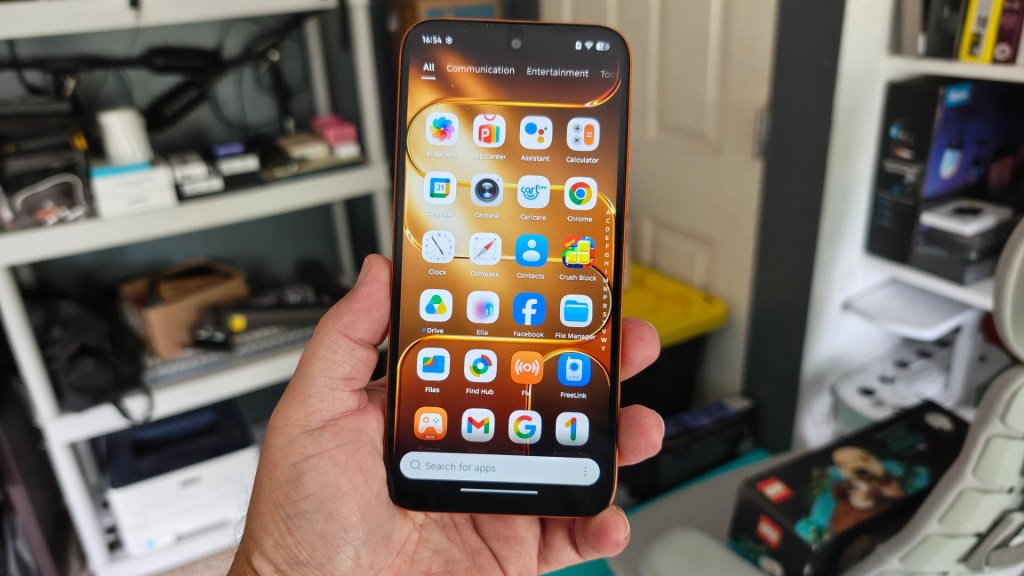 A person holding a smartphone displaying various app icons on the screen, with a background of shelves containing electronic items and accessories.