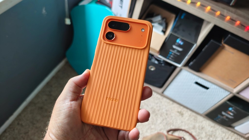 A hand holding an orange smartphone with a textured case, featuring dual rear cameras and the brand name 'Tecno' visible.