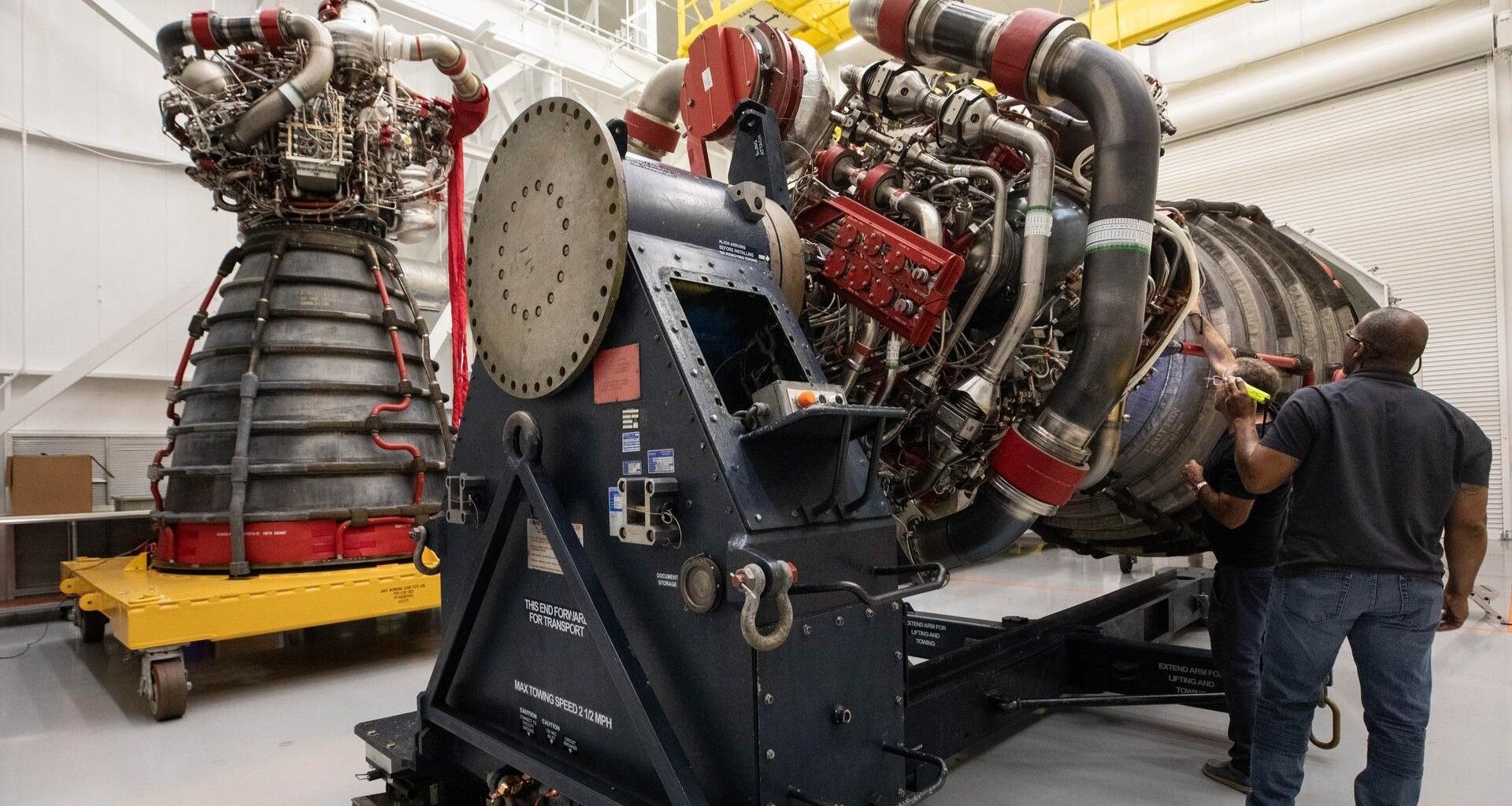 Two large rocket engines, one standing, one horizontal, rest on support platforms inside a white industrial setting.