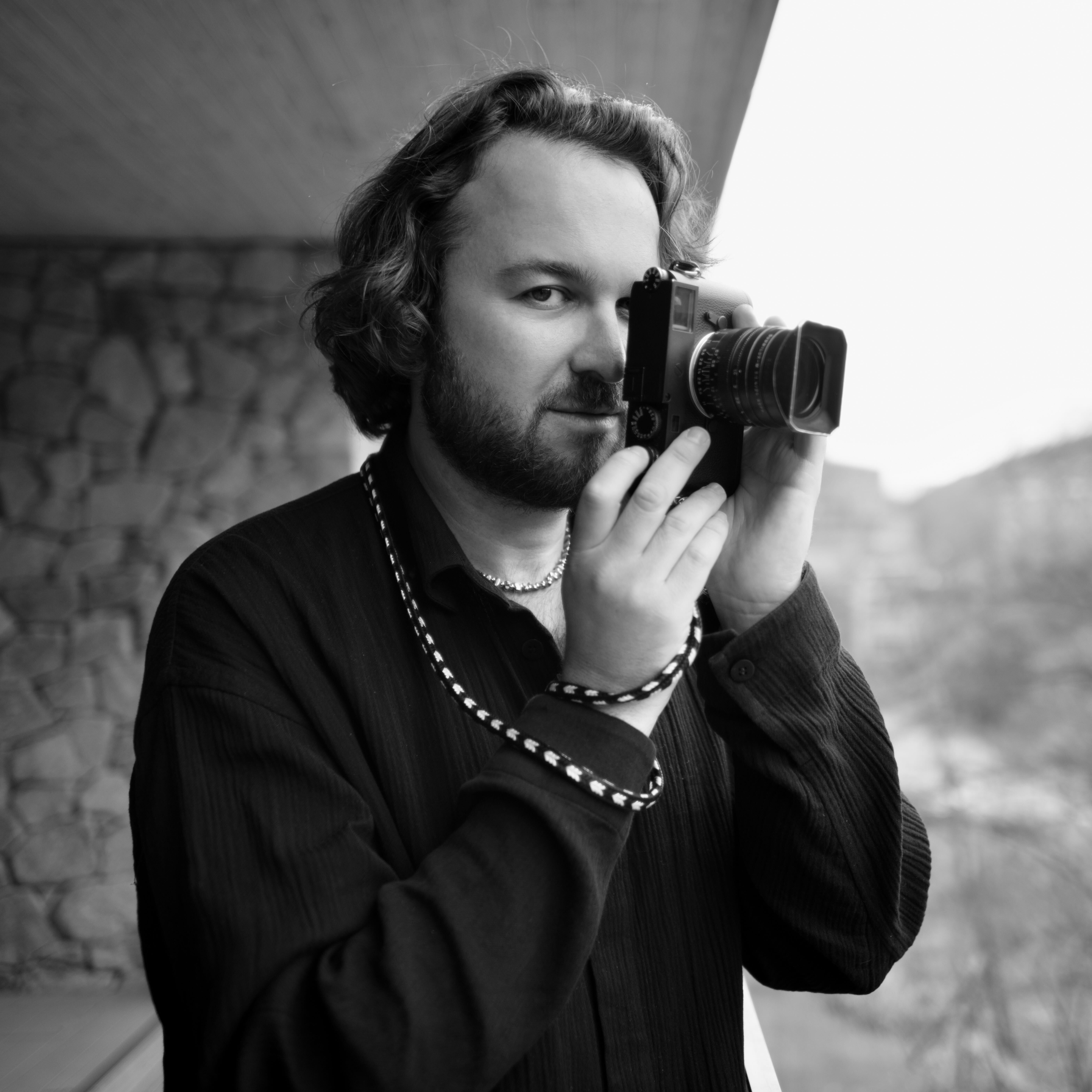A black and white portrait of Alan Schaller holding a Leica camera up to his eye, photographed by Ryota Kohama.