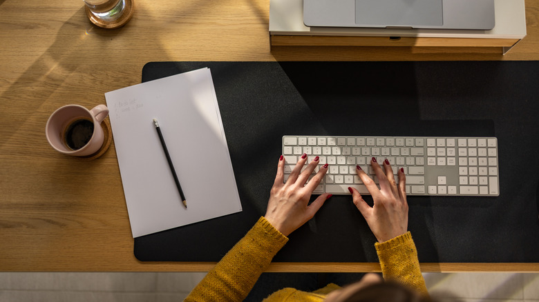 A top-down view of a person typing on a white wireless keyboard next to a notebook and a coffee mug.